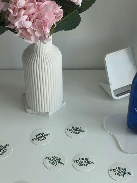 White vase with pink flowers on a white surface with 'High Standards Only' stickers.